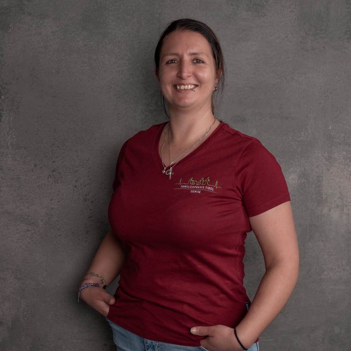 Denise, cleaning, hygiene and sterilisation assistant at Familienpraxis Tirol, in a burgundy polo shirt with the practice logo, smiling against a grey background.