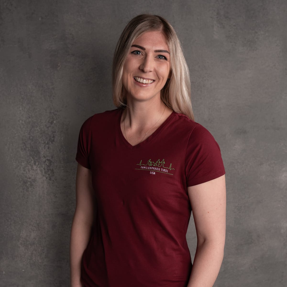 Lisa, registered nurse (DGKP) at Familienpraxis Tirol, in a burgundy polo shirt with the practice logo, smiling against a grey background.
