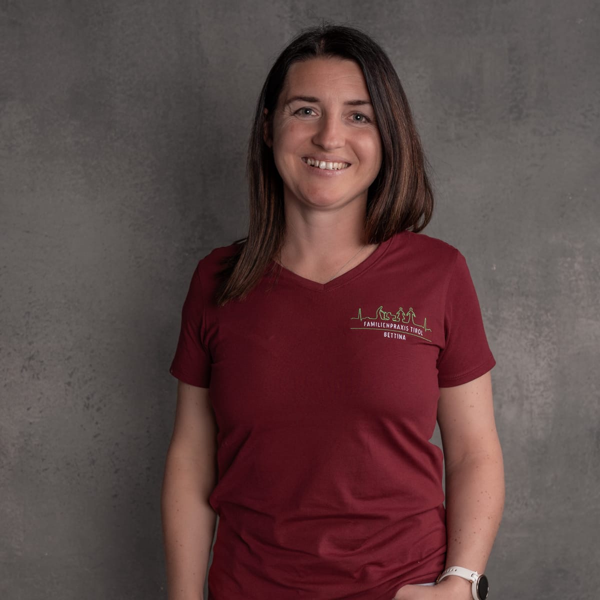Bettina, practice assistant at Familienpraxis Tirol, in a burgundy polo shirt with the practice logo, smiling against a grey background.