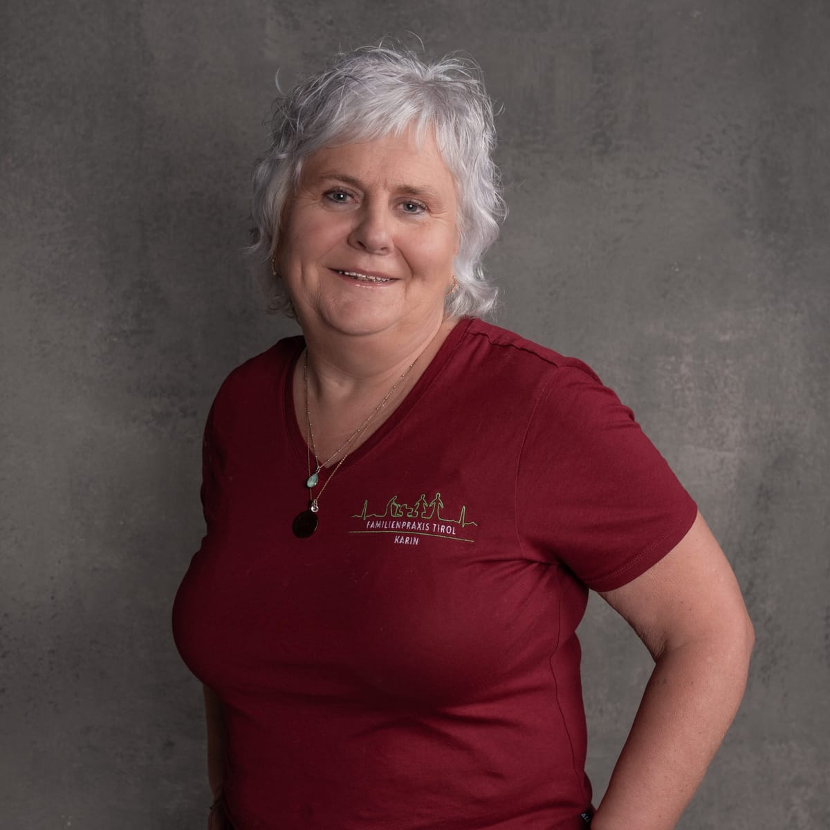 Karin, practice assistant at Familienpraxis Tirol, in a burgundy polo shirt with the practice logo, smiling against a grey background.