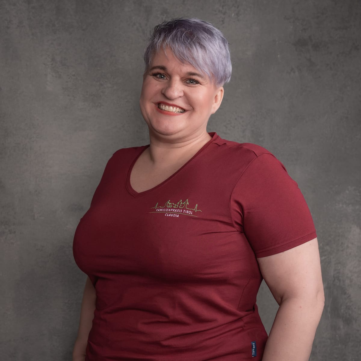 Claudia, nursing assistant at Familienpraxis Tirol, in a burgundy polo shirt with the practice logo, smiling against a grey background.