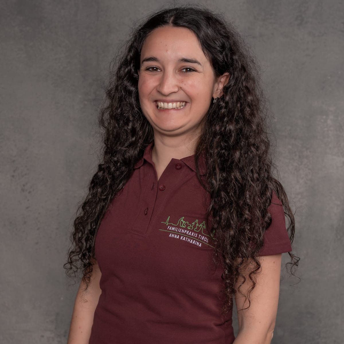 Anna Katharina, practice manager at Familienpraxis Tirol, in a burgundy polo shirt with the practice logo, smiling against a grey background.