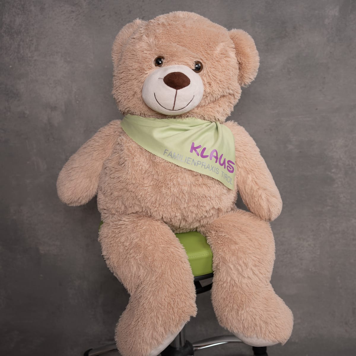 Klaus, the mascot of Familienpraxis Tirol – a large beige teddy bear with a neckerchief and the practice logo, sitting on a green office chair against a grey background.